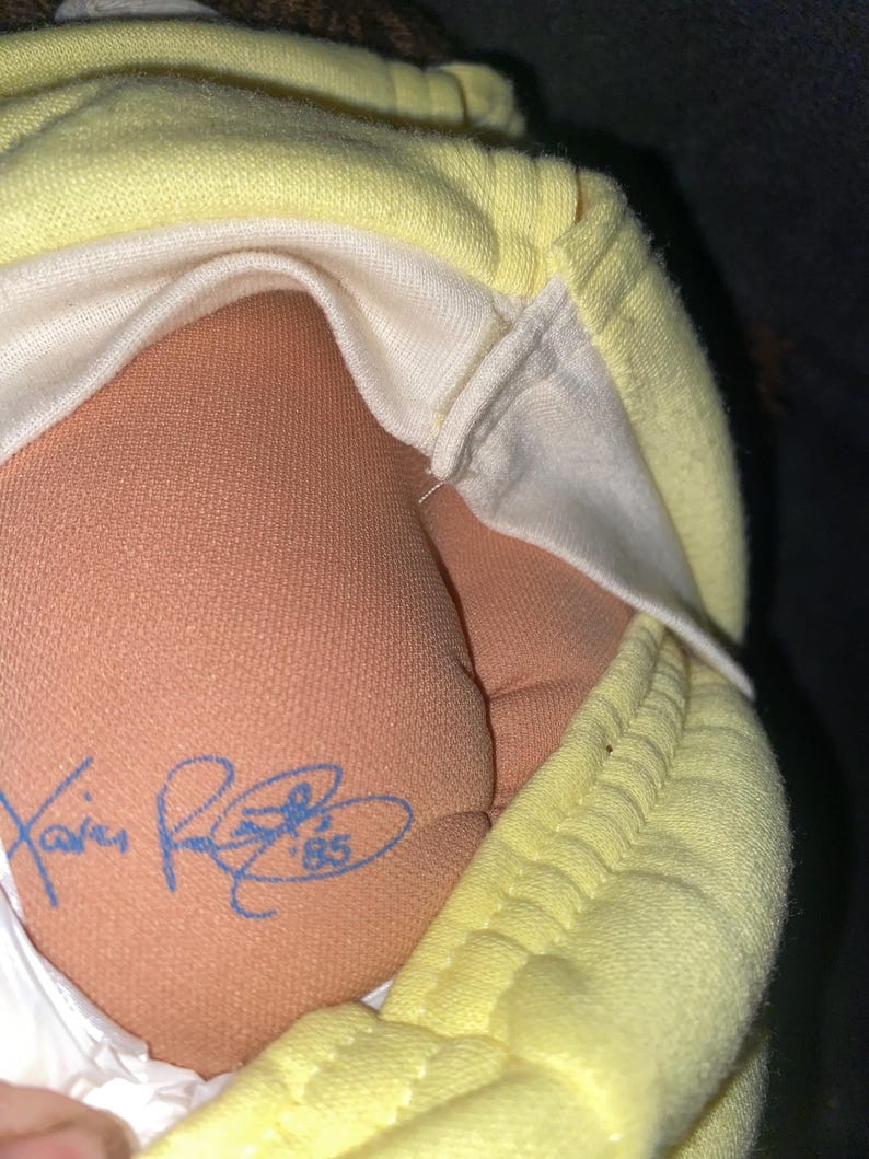 Op de afbeelding: Een pop met een huid van beige stof die een lichtgeel kledingstuk draagt. De pop heeft een blauwe inkt-handtekening op de onderrug met de tekst Xavier Roberts '85. De stof van het kledingstuk is gestructureerd en lichtgeel van kleur.
