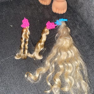 Puede incluir: Tres mechones de pelo rubio rizado con pinzas rosas y azules. El pelo est&aacute; separado en dos mechones m&aacute;s peque&ntilde;os y uno m&aacute;s grande.