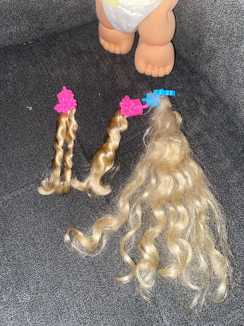 Puede incluir: Tres mechones de pelo rubio rizado con pinzas rosas y azules. El pelo est&aacute; separado en dos mechones m&aacute;s peque&ntilde;os y uno m&aacute;s grande.