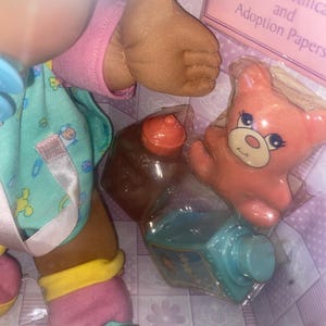 Pu&ograve; includere: Una bambola dalla pelle scura indossa una maglietta color verde acqua con motivi colorati, maniche rosa e calzini rosa e gialli. Gli accessori includono un piccolo orso arancione, una bottiglia marrone e una bottiglia verde acqua in una scatola con la scritta Birth Certificate and Adoption Papers.