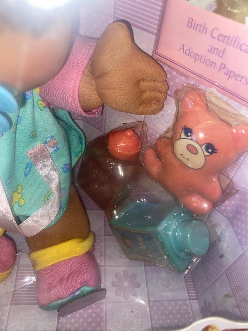 Pu&ograve; includere: Una bambola dalla pelle scura indossa una maglietta color verde acqua con motivi colorati, maniche rosa e calzini rosa e gialli. Gli accessori includono un piccolo orso arancione, una bottiglia marrone e una bottiglia verde acqua in una scatola con la scritta Birth Certificate and Adoption Papers.
