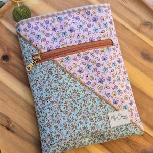 May include: A floral print pouch with a brown zip and gold-coloured zip pull. The pouch features a patchwork design with light blue and pink fabric, adorned with butterfly and flower patterns. The pouch has a small tag with the text "Ma Cherie".