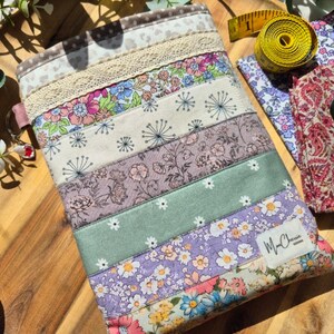 May include: A patchwork fabric case featuring floral patterns in pink, blue, and purple. The case is constructed from horizontal fabric strips, some with lace trim. The label "Ma Cherie Designs" is visible on the bottom right.
