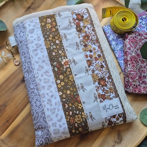May include: A patchwork fabric case with floral, bird, and animal print panels in brown, cream, and grey. The case has a cream lace trim. A yellow tape measure is visible in the background. The case is likely for storing items.