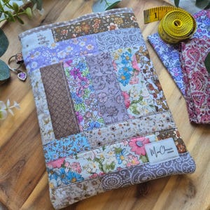 May include: A patchwork tablet sleeve in a variety of floral and patterned fabrics, featuring shades of brown, purple, and blue. The sleeve is rectangular and has a label reading "McCherie Designs". A yellow tape measure and fabric pieces are in the background.