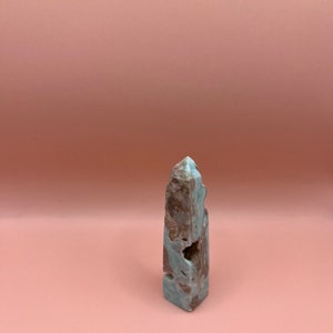 May include: A natural, rectangular crystal tower with a light blue and brown color scheme. The crystal has a rough, textured surface with a pointed top and a hollowed-out section on one side. The crystal is standing on a pink background.