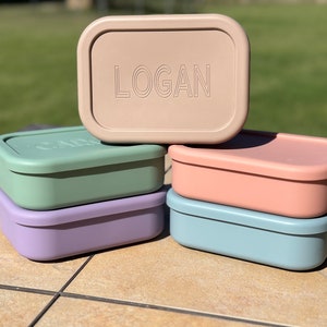 May include: Stack of five rectangular lunch boxes in various colors. The top box is beige with the name "LOGAN" embossed on the lid. The other boxes are green, pink, light blue, and purple.