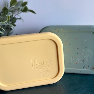 May include: Two yellow and green silicone lunchboxes with the names "Rosie" and "Peter" embossed on the lids. Each lid has a small animal design.