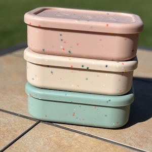 May include: Three stackable lunch containers with speckled lids. The top container is pink with white and multi-colored speckles. The middle container is white with yellow and multi-colored speckles. The bottom container is light blue with orange and multi-colored speckles.