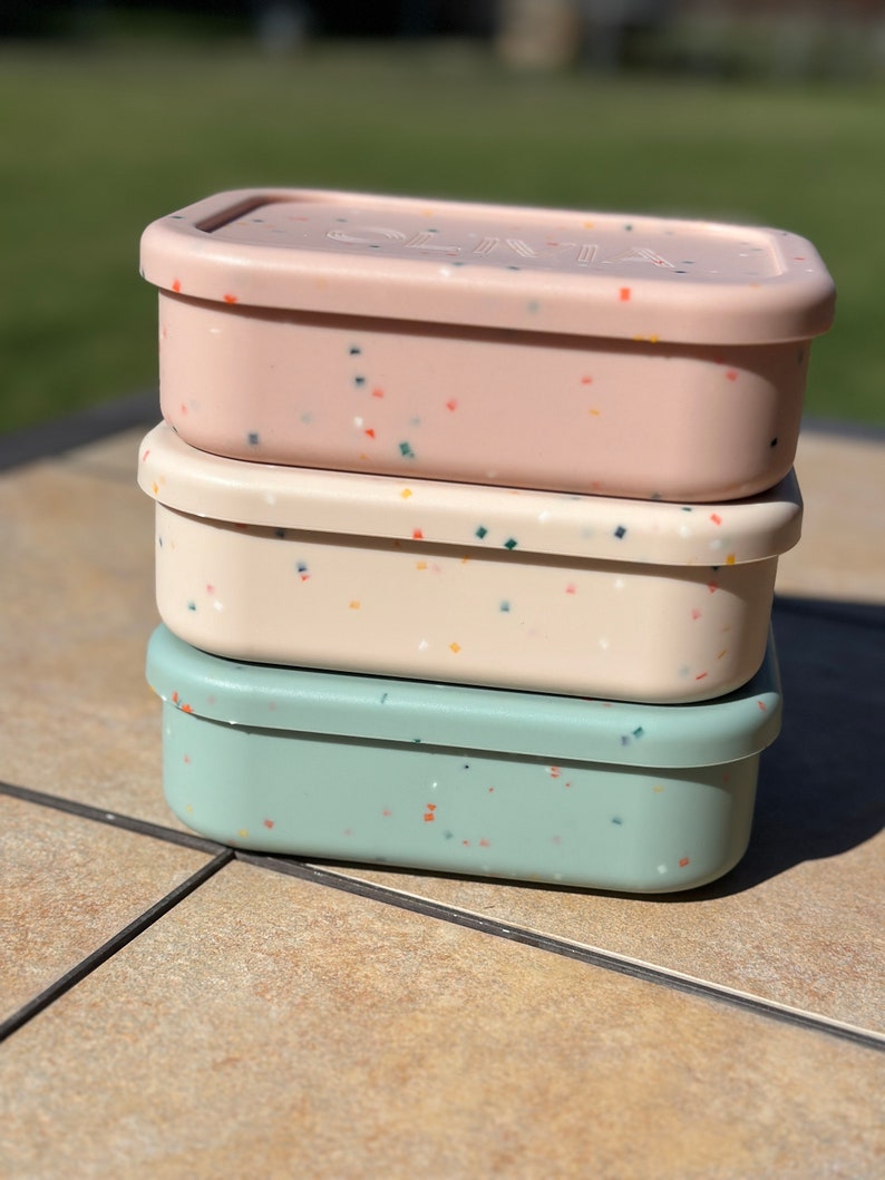 May include: Three stackable lunch containers with speckled lids. The top container is pink with white and multi-colored speckles. The middle container is white with yellow and multi-colored speckles. The bottom container is light blue with orange and multi-colored speckles.
