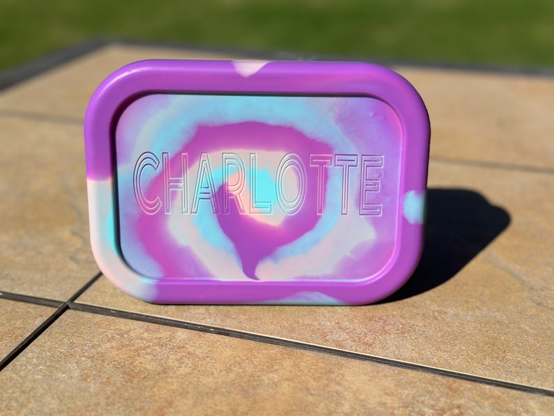 May include: A purple rectangular container with a swirl design in shades of blue, pink, and white. The name "CHARLOTTE" is embossed in white on the container.