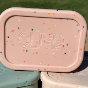 May include: A pink rectangular lunchbox with a speckled pattern and the name "OLIVIA" embossed on the front.