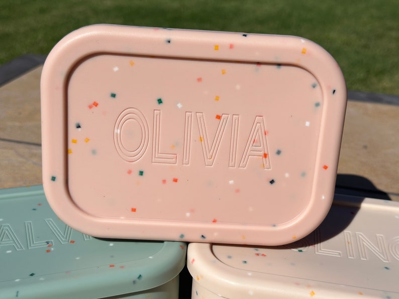 May include: A pink rectangular lunchbox with a speckled pattern and the name "OLIVIA" embossed on the front.