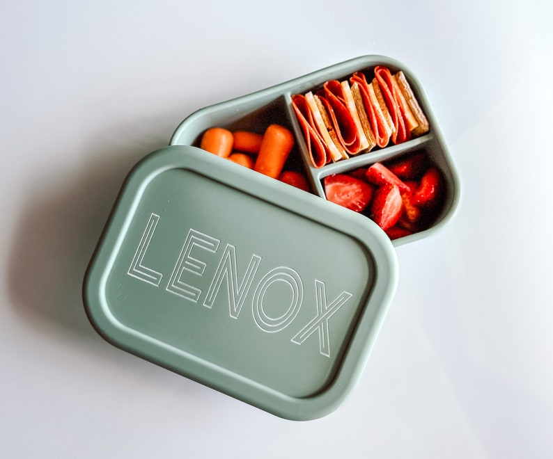 May include: A green silicone lunch box with the name "LENOX" printed on the lid. The box is open and contains a variety of snacks, including strawberries, carrots, and sliced meat.