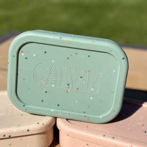 May include: A green lunchbox with a speckled pattern and the name "CALVIN" printed on the lid.
