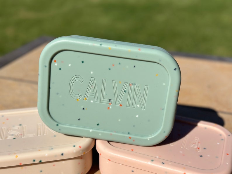 May include: A green lunchbox with a speckled pattern and the name "CALVIN" printed on the lid.
