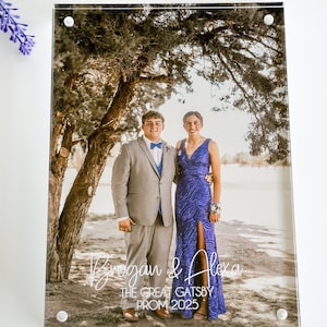 May include: A clear acrylic photo frame with a photograph of a couple in formal attire. The photo is of a man in a gray suit and a woman in a blue gown. The frame has the text "Brogan & Alexa" and "The Great Gatsby PROM 2025".