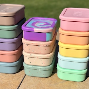 May include: A stack of six colorful silicone food storage containers with a speckled pattern, and a stack of five silicone food storage containers with a solid color. The containers are all square shaped and have a lid.