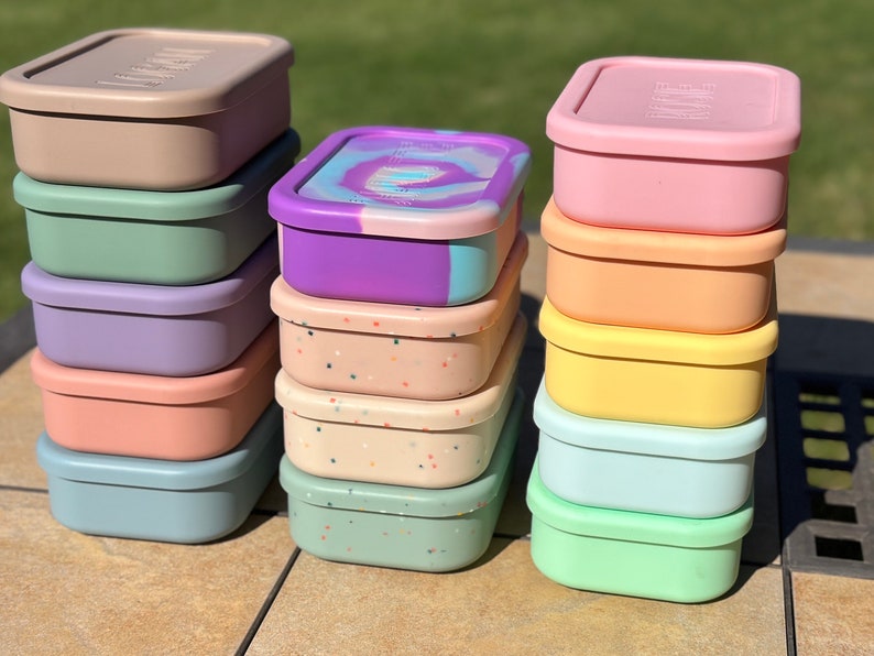 May include: A stack of six colorful silicone food storage containers with a speckled pattern, and a stack of five silicone food storage containers with a solid color. The containers are all square shaped and have a lid.