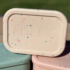 May include: A pale pink rectangular container with a speckled pattern and the word "AISLING" embossed on the top.