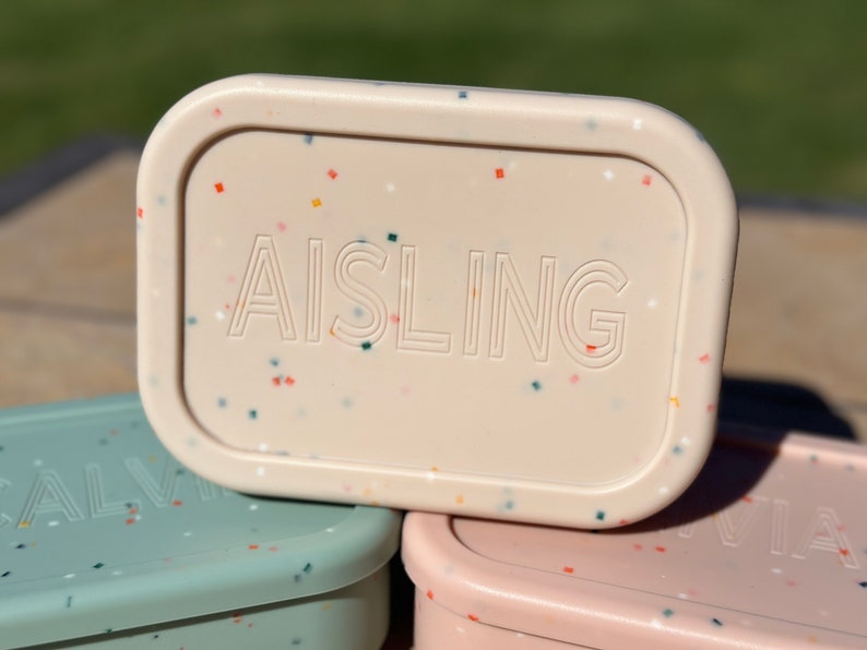 May include: A pale pink rectangular container with a speckled pattern and the word "AISLING" embossed on the top.