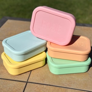 May include: Stack of five pastel colored rectangular lunch boxes. The top box is pink with the name "ROSIE" embossed on the lid. The next box is light blue, followed by yellow, green, and orange.