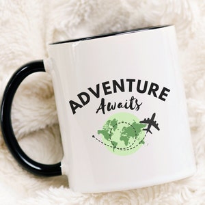 May include: White ceramic mug with a black handle and rim. The mug has a green and black graphic of a world map with a plane and the text "Adventure Awaits".