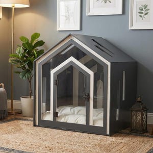May include: A dark gray pet house with a gabled roof, white trim, and a clear front panel door. The structure features a hinged door with a black latch and contains a white cushioned interior floor, providing a contained space for a pet.