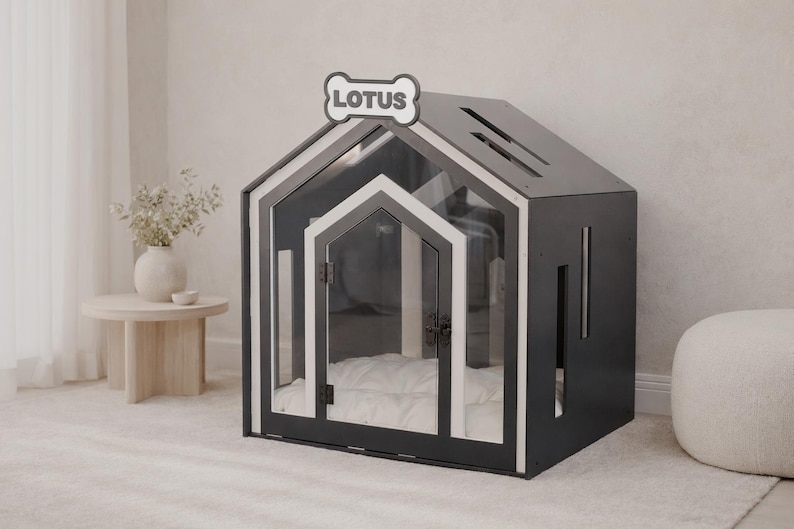 May include: A dark gray and white pet house with a gabled roof and a clear front door. The structure features a bone-shaped sign at the peak with the text LOTUS printed in black. Inside, a white cushioned bed is visible through the transparent door.