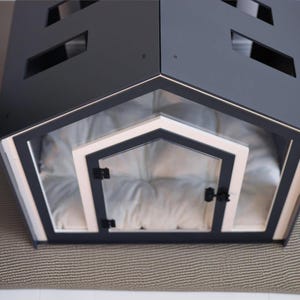 May include: A dark gray pet house with a gabled roof featuring rectangular cutouts. The front entrance has a light-colored frame and a clear door with black hinges and a latch. Inside, a soft, tufted white cushion fills the floor space.