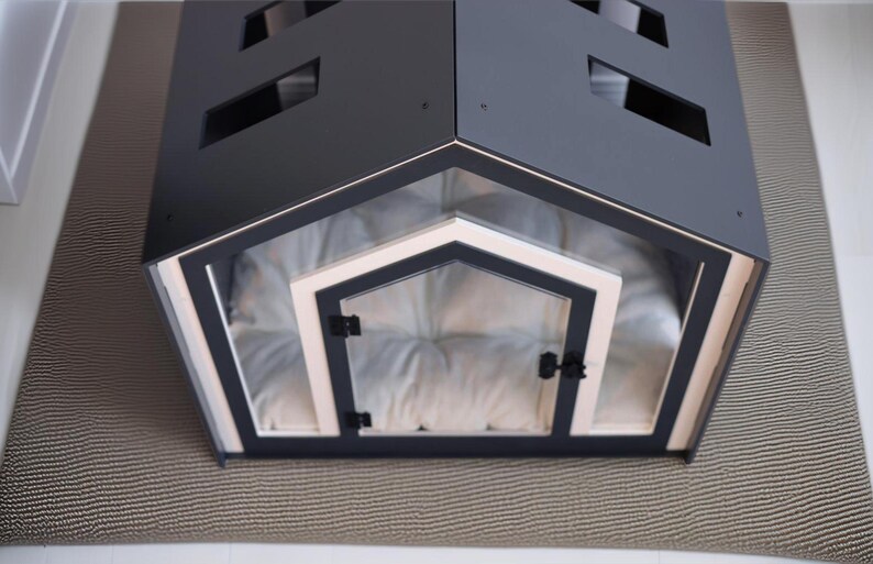 May include: A dark gray pet house with a gabled roof featuring rectangular cutouts. The front entrance has a light-colored frame and a clear door with black hinges and a latch. Inside, a soft, tufted white cushion fills the floor space.