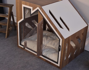May include: A wooden pet house with a dark wood grain finish and a white roof panel. The structure features a house-shaped front frame, a clear acrylic door with metal hinges, and a white tufted cushion inside. Geometric cutouts decorate the side.