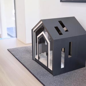 May include: A dark gray pet house shaped like a small cabin with a white-trimmed arched entrance. The structure features rectangular cutouts on the roof and side walls, and a soft white cushion is visible inside the interior space.