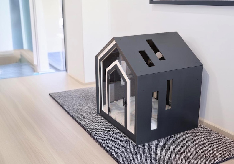 May include: A dark gray pet house shaped like a small cabin with a white-trimmed arched entrance. The structure features rectangular cutouts on the roof and side walls, and a soft white cushion is visible inside the interior space.