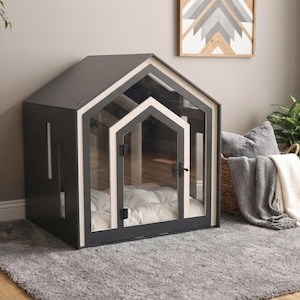 May include: A dark gray pet house with a gabled roof and a clear acrylic front panel featuring a white frame. The structure includes a hinged door with a black latch and a white tufted cushion inside for comfort, creating a secure enclosure.