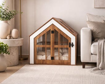 Puede incluir: Una casa para mascotas de madera con techo a dos aguas, con un marco blanco y dos puertas con paneles de vidrio, bisagras de metal negro y un pestillo. Dentro de la estructura, se puede ver un cojín blanco suave y mullido.