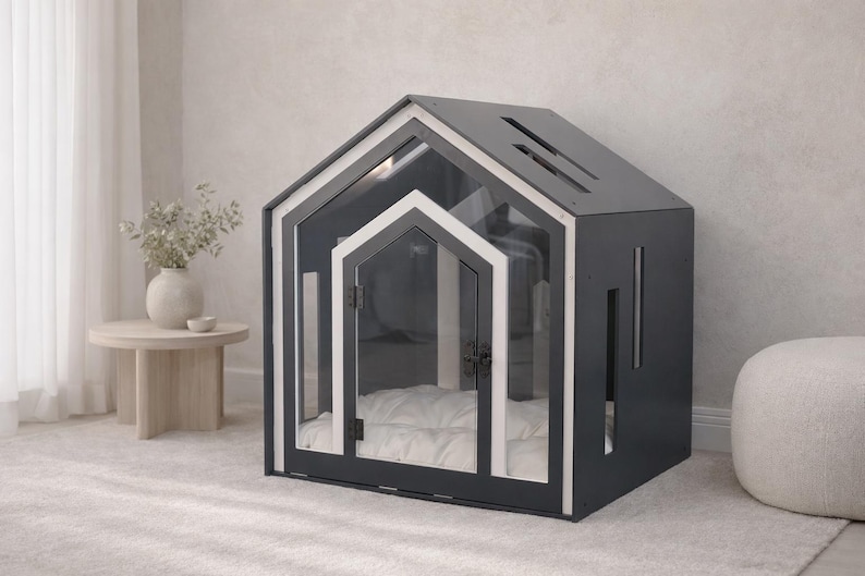 May include: A dark gray pet house with a gabled roof, featuring a clear acrylic front panel, a white-trimmed door frame, and a soft white tufted cushion inside. The structure includes vertical side cutouts and a slanted roof with rectangular vents.