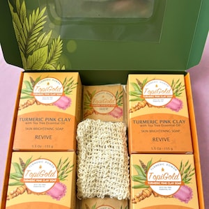 May include: An open green box filled with orange boxes of TopiGold Turmeric Pink Clay Soap, a small soap, and a beige washcloth. The boxes have text and illustrations of turmeric and pink clay. The soap is 5.5 oz (155 g).