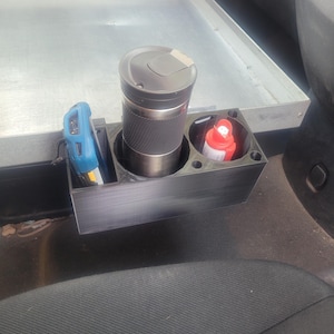 May include: A black 3D-printed car cup holder with three compartments. One holds a black and silver travel mug, another a red and white bottle, and the third a blue tool. The holder sits on a car seat.