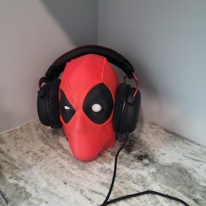 May include: A red Deadpool mask with black eyes and a white mouth is wearing black headphones with red accents. The headphones are connected to a black cord.