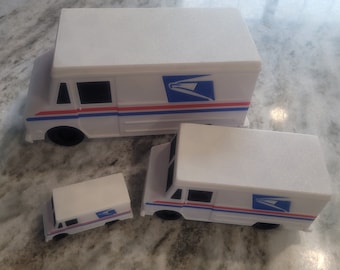 May include: Three white toy mail trucks of different sizes. The largest truck has a blue and red stripe and the USPS logo. The medium truck has a blue and red stripe and the USPS logo. The smallest truck has a blue and red stripe and the USPS logo.