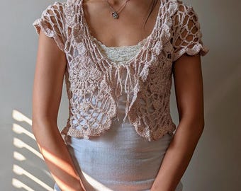 May include: A delicate, crocheted, blush-colored cropped cardigan with a tie front. The cardigan has a lacy, open-weave design and short sleeves.