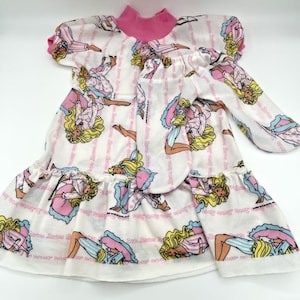 May include: White doll dress with a pink collar and cuffs, featuring a repeating pattern of a blonde doll in pajamas. The dress has a ruffled skirt and long sleeves. The doll's name is printed in pink on the dress.
