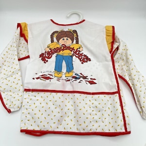 May include: White Cabbage Patch Kids apron with long sleeves. The apron has a red trim and a graphic of a Cabbage Patch Kids doll. The sleeves have a pattern of small yellow flowers. The apron is hanging on a white hanger.