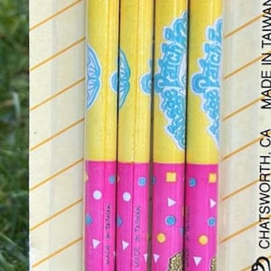 May include: A pack of Cabbage Patch Kids pencils. The pencils are pink and yellow with a repeating pattern of the Cabbage Patch Kids logo and characters. The pencils have gold-coloured metal bands and white erasers.
