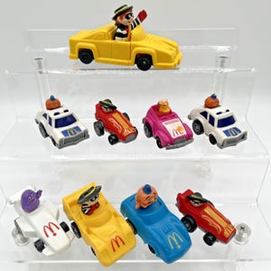 May include: A selection of vintage McDonald's Happy Meal toys. The toys feature characters in toy cars of various colours, including yellow, red, pink, blue, and white. Characters include Hamburglar and Officer Big Mac.