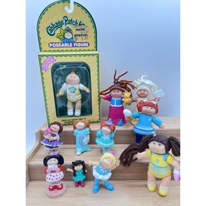Peut inclure: Une collection de figurines articulées Cabbage Patch Kids, dont une dans son emballage d'origine jaune et vert. Les figurines présentent diverses tenues, couleurs de cheveux et poses, certaines tenant des accessoires comme un ours en peluche.