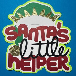 May include: A layered paper cutout sign featuring the text "Santa's little HELPER" in red and black lettering. The design includes a green elf hat with a white trim and gold glitter accents, all set against a bright neon green border.
