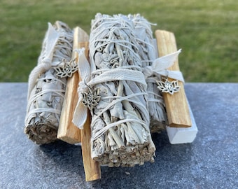 May include: Three bundles of white sage smudge sticks tied with white string and decorated with silver charms. The smudge sticks are arranged on a wooden base.