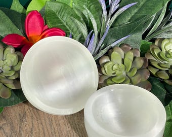 May include: Two white selenite bowls, one slightly larger than the other, are shown on a wooden surface. The bowls are smooth and have a subtle shimmer. The bowls are surrounded by green foliage.
